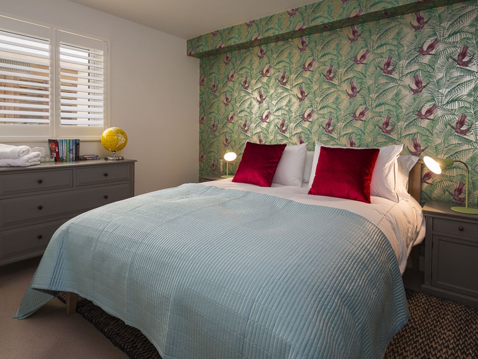 A bedroom with a double bed with red pillows and a blue bedspread next to a grey dresser and nightstand at Villa 8 Estura Salcombe