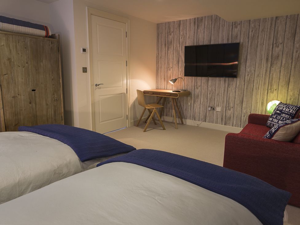 A bedroom with two beds a wooden wardrobe a desk with a chair and lamp a wall mounted TV and a red sofa at Villa 8 Estura in Salcombe